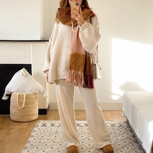 May include: A woman wearing a white sweater, white pants, and brown suede boots. She is wearing a plaid scarf with pink, brown, and burgundy colors. She is holding a white phone in her hand.