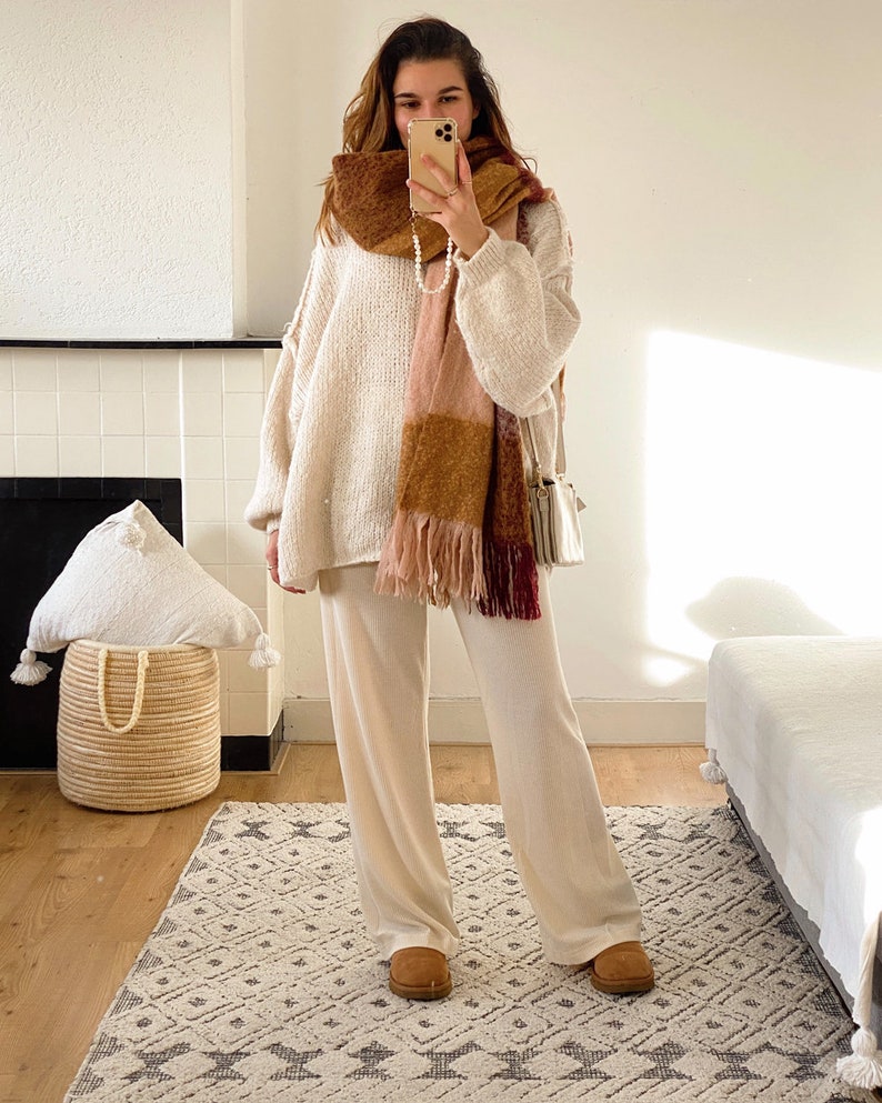 May include: A woman wearing a white sweater, white pants, and brown suede boots. She is wearing a plaid scarf with pink, brown, and burgundy colors. She is holding a white phone in her hand.
