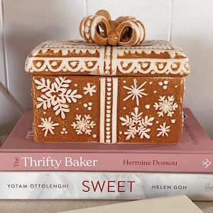 Op de afbeelding: Een bruine keramische geschenkdoos met witte sneeuwvlokken en decoratieve randdetails. Het deksel heeft een strikvormige handgreep. De doos staat op twee boeken, waarvan er één het woord "SWEET" draagt.