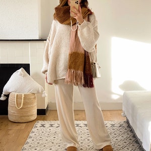 May include: A woman wearing a white knit sweater and matching pants with a brown, pink, and burgundy plaid scarf. She is standing on a patterned rug in front of a fireplace.