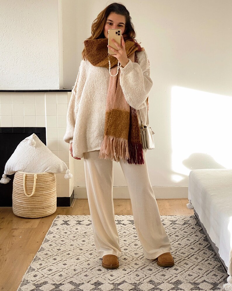May include: A woman wearing a white knit sweater and matching pants with a brown, pink, and burgundy plaid scarf. She is standing on a patterned rug in front of a fireplace.