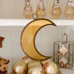 Può includere: Un ornamento a forma di luna crescente dorato con una superficie a specchio. L'ornamento è appeso a una corda dorata ed è circondato da ornamenti natalizi bianchi e dorati.