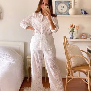 May include: A white eyelet  shirt and pants set. The shirt has a button-up front and long sleeves. The pants are wide-leg and have a relaxed fit.