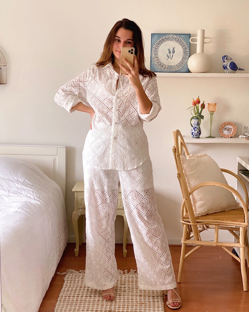 May include: A white eyelet  shirt and pants set. The shirt has a button-up front and long sleeves. The pants are wide-leg and have a relaxed fit.