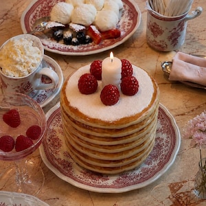 May include: A tall stack of golden-brown pancakes topped with fresh red raspberries, a dusting of white powdered sugar, and a single lit white candle. The pancakes are served on a white plate with a red floral pattern, surrounded by other dishes.