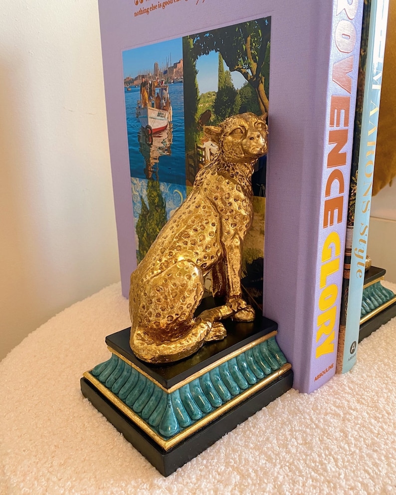 K&ouml;nnte beinhalten: Ein goldfarbener Geparden-Buchst&uuml;tze zwischen zwei B&uuml;chern. Die Buchst&uuml;tze steht auf einem schwarzen Sockel mit t&uuml;rkisfarbenen und goldenen Akzenten. Der Gepard ist mit Flecken und einer k&ouml;niglichen Pose versehen. Die B&uuml;cher haben farbenfrohe Einb&auml;nde.