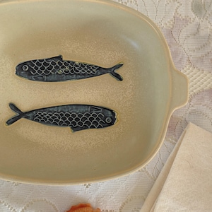 May include: Cream-colored ceramic serving dish with two stylized blue fish designs. The oval dish has a slightly raised rim and small handles. A seashell and a folded linen napkin are also visible.