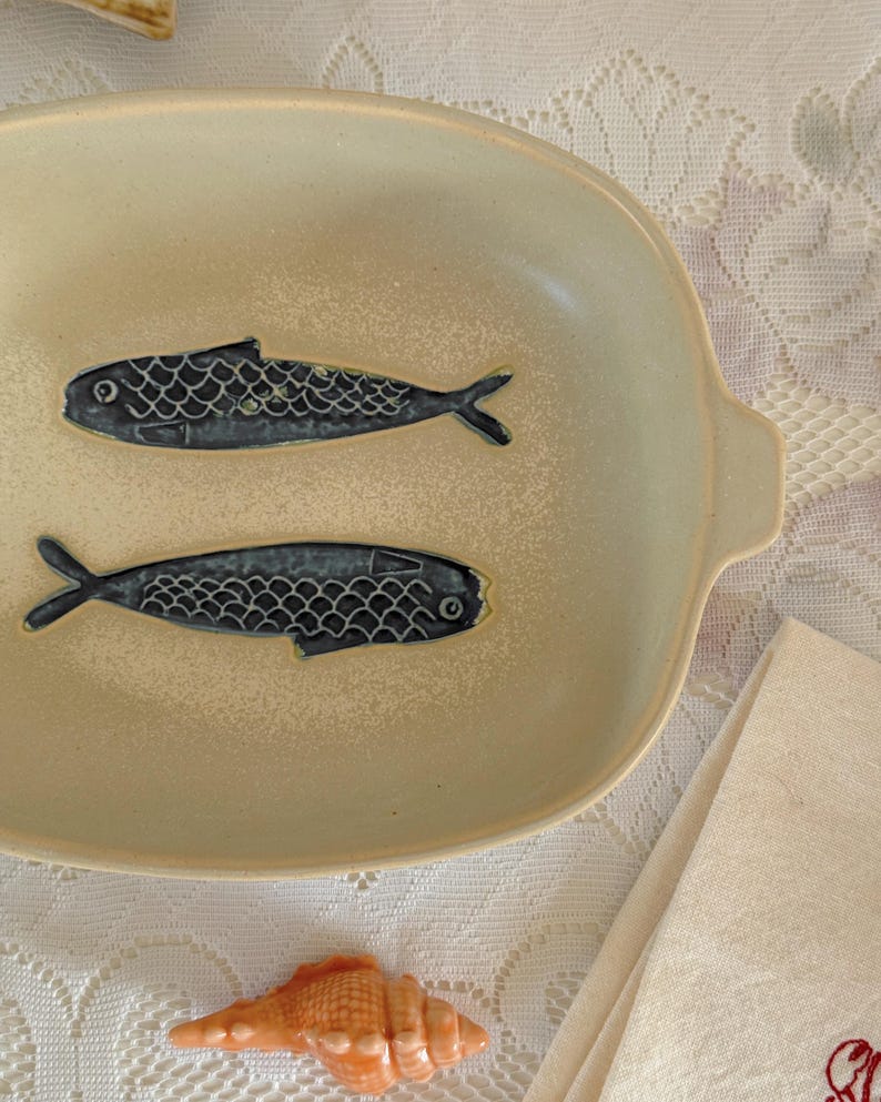 May include: Cream-colored ceramic serving dish with two stylized blue fish designs. The oval dish has a slightly raised rim and small handles. A seashell and a folded linen napkin are also visible.