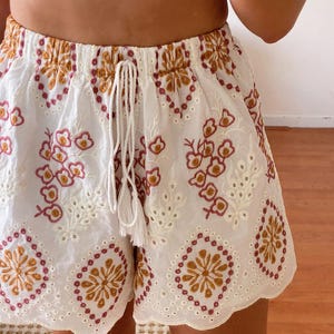 K&ouml;nnte beinhalten: Wei&szlig;e H&auml;kelshorts mit einem gewellten Saum und einem gestickten Blumenmuster in Braun- und Rosat&ouml;nen.