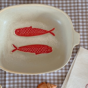 May include: Cream-colored ceramic serving dish with two red fish illustrations. The dish has a rectangular shape with rounded corners and small handles. The fish are detailed with scales and fins.