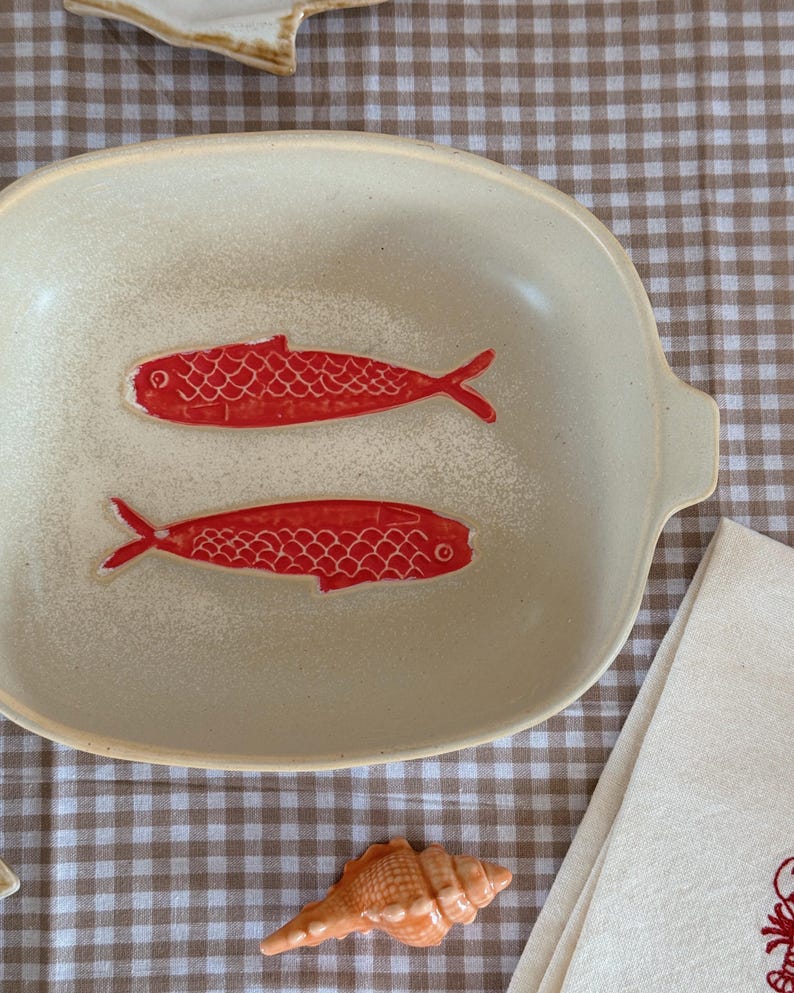 May include: Cream-colored ceramic serving dish with two red fish illustrations. The dish has a rectangular shape with rounded corners and small handles. The fish are detailed with scales and fins.