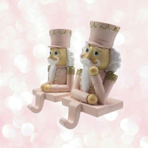 May include: Two pink and gold nutcracker stocking holders. The nutcrackers are wearing pink and gold uniforms and have white beards and moustaches. They are sitting on a pink base with a gold hook.