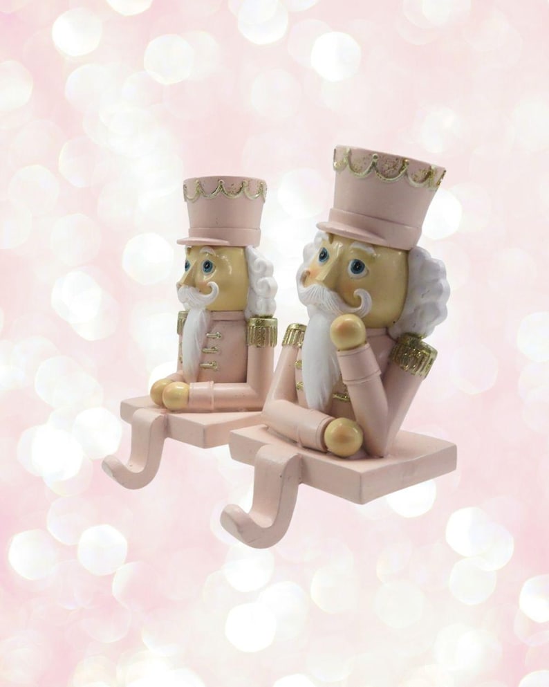 May include: Two pink and gold nutcracker stocking holders. The nutcrackers are wearing pink and gold uniforms and have white beards and moustaches. They are sitting on a pink base with a gold hook.