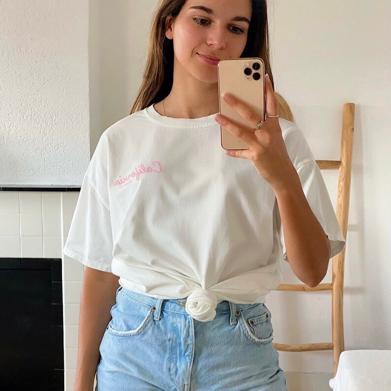 K&ouml;nnte beinhalten: Ein wei&szlig;es T-Shirt mit einem rosa gestickten Design, auf dem vorne "California" steht. Das Shirt ist in der Taille gebunden und zu einer hellblauen Jeans-Shorts kombiniert.