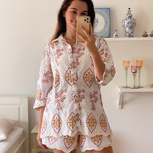 K&ouml;nnte beinhalten: Ein wei&szlig;es Spitzenoberteil und -shorts-Set mit einem braunen und rosafarbenen Blumenmuster. Das Oberteil hat einen Knopfleisten vorne und Dreiviertel&auml;rmel. Die Shorts haben einen gewellten Saum.