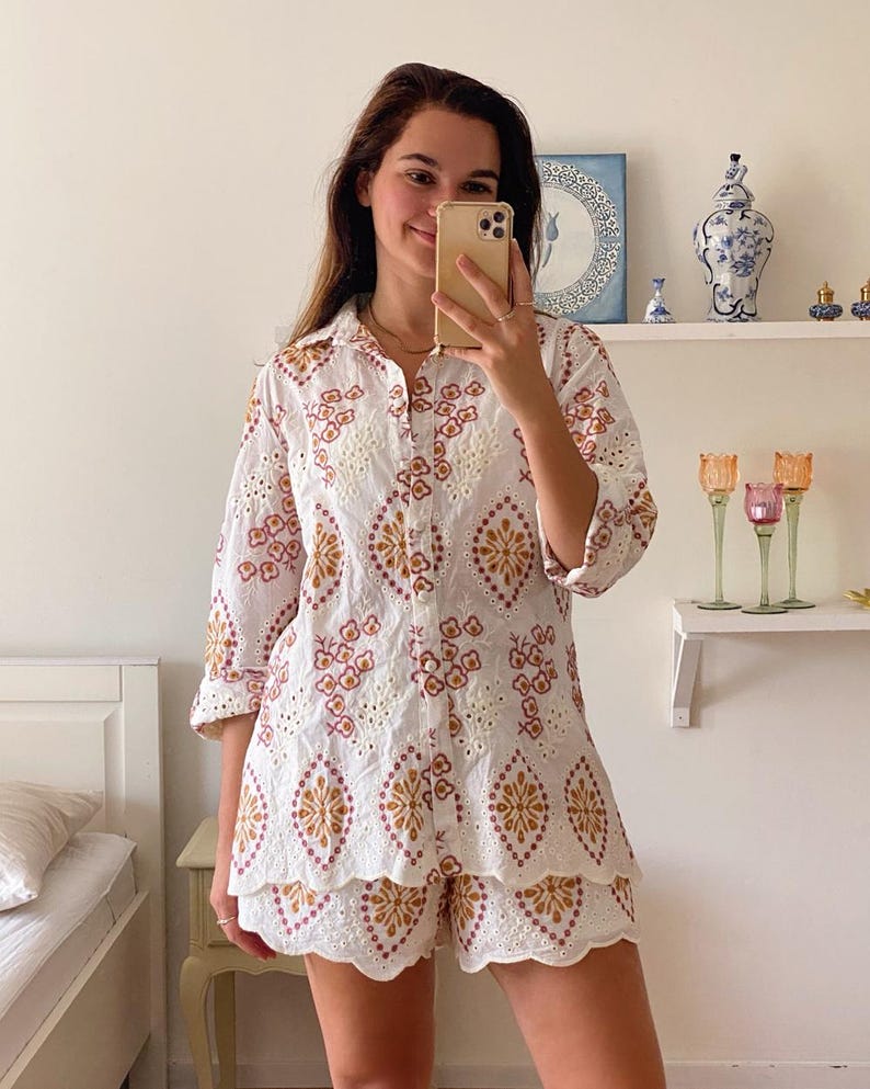 K&ouml;nnte beinhalten: Ein wei&szlig;es Spitzenoberteil und -shorts-Set mit einem braunen und rosafarbenen Blumenmuster. Das Oberteil hat einen Knopfleisten vorne und Dreiviertel&auml;rmel. Die Shorts haben einen gewellten Saum.