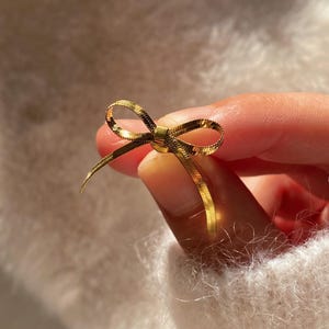 May include: A gold-colored bow-shaped jewelry piece. The bow is made of a thin, woven material, and the ends of the bow are slightly curved. The jewelry is held between fingers against a soft, white background.
