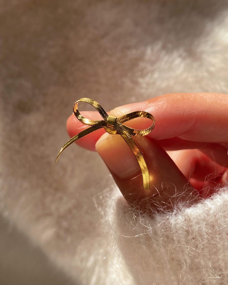 May include: A gold-colored bow-shaped jewelry piece. The bow is made of a thin, woven material, and the ends of the bow are slightly curved. The jewelry is held between fingers against a soft, white background.