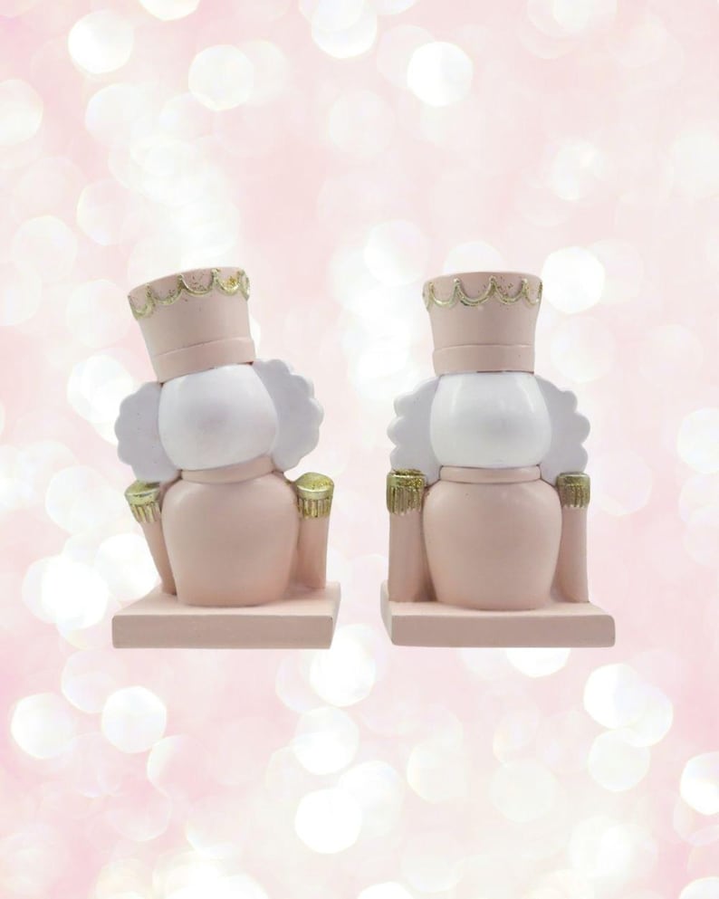 May include: Two pink and white nutcracker figurines with gold accents. The nutcrackers are standing on a pink base.