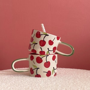 May include: Two white ceramic mugs with green handles stacked on top of each other. Both mugs have a red cherry pattern.