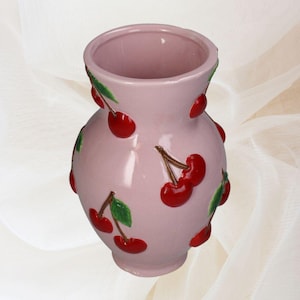 Op de afbeelding: Een roze keramische vaas met een glanzende afwerking. De vaas is versierd met verhoogde rode kersendesigns en groene bladeren. De vaas heeft een afgeronde body en een licht uitlopende opening, ideaal voor bloemen of als decoratief item.