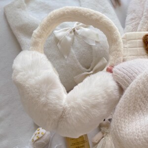 May include: Ivory-colored earmuffs with a fluffy, faux fur texture. The headband is also covered in the same material. A small, white bow adorns the center of the earmuffs. The image suggests a cozy, winter accessory.