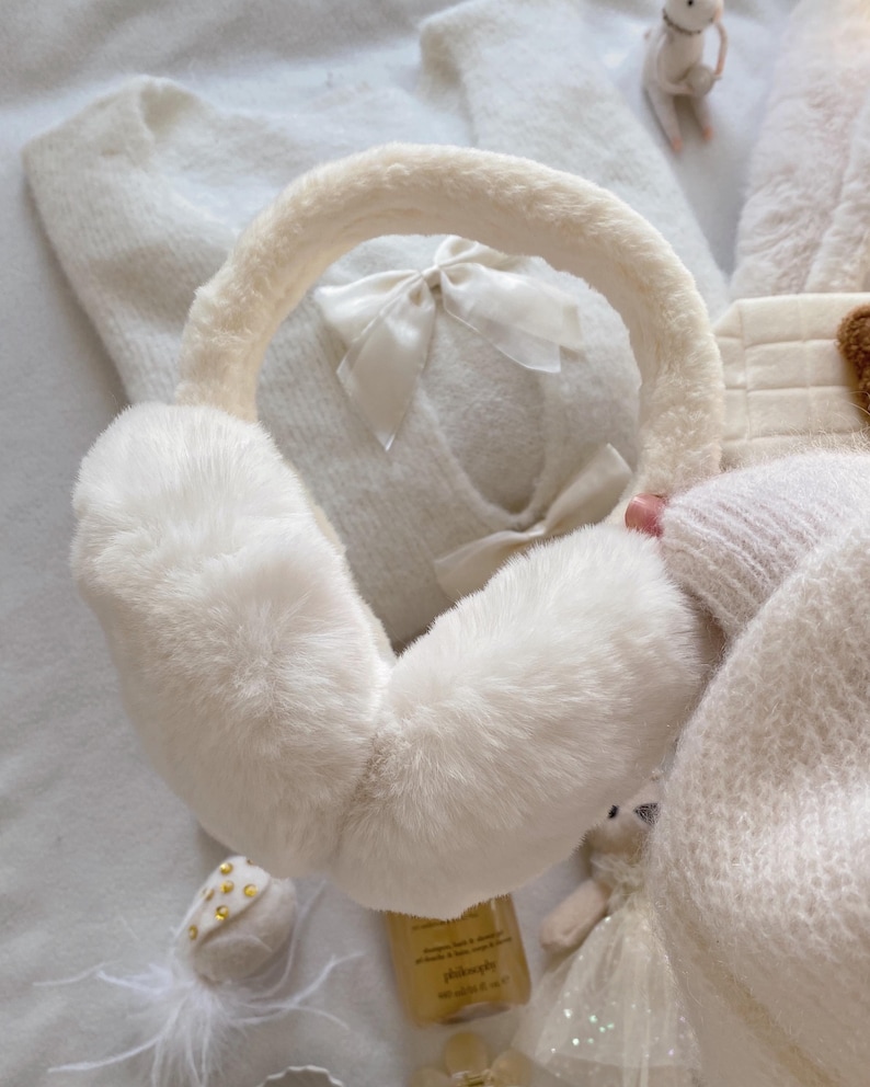 May include: Ivory-colored earmuffs with a fluffy, faux fur texture. The headband is also covered in the same material. A small, white bow adorns the center of the earmuffs. The image suggests a cozy, winter accessory.