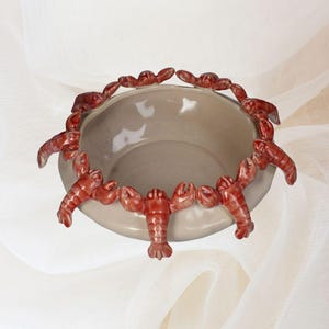 May include: A ceramic bowl with a light gray interior and a rim decorated with red lobster figurines. The lobsters are arranged around the edge of the bowl, creating a unique and decorative design. The bowl is likely used for serving or display.