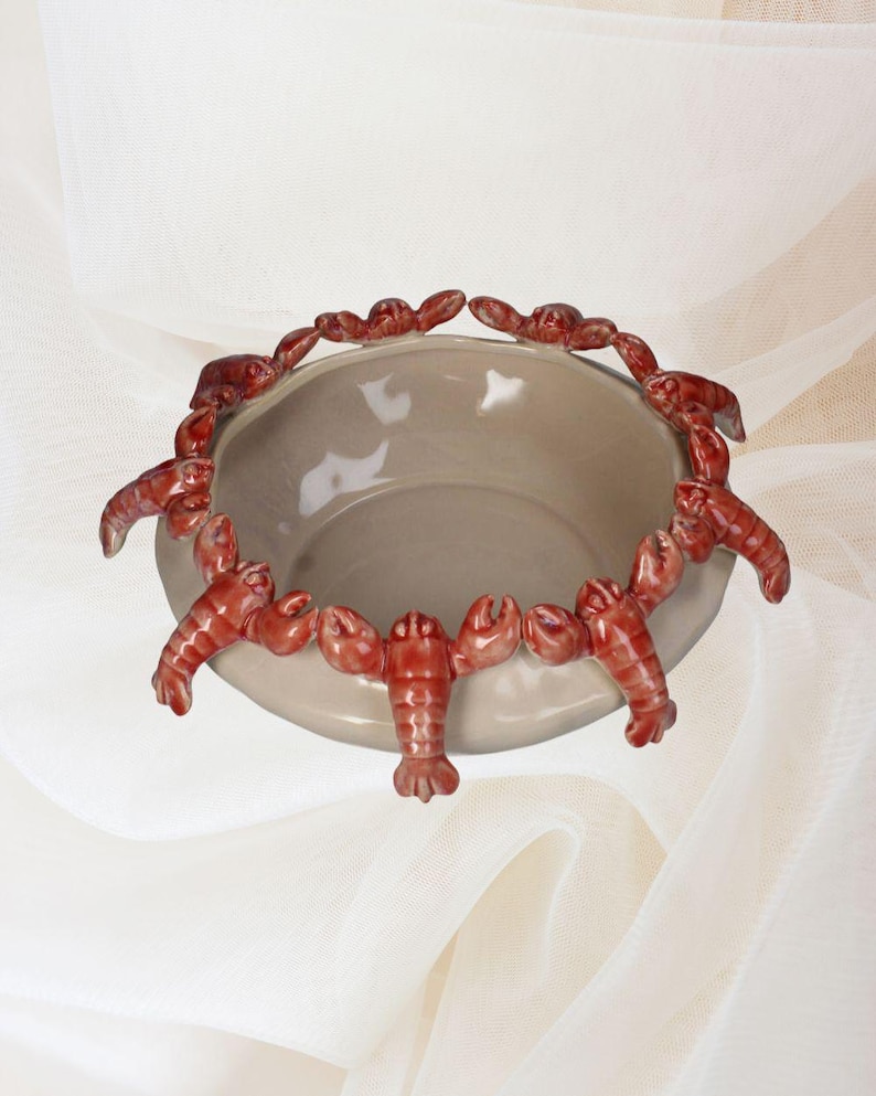 May include: A ceramic bowl with a light gray interior and a rim decorated with red lobster figurines. The lobsters are arranged around the edge of the bowl, creating a unique and decorative design. The bowl is likely used for serving or display.