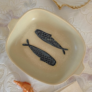 May include: A beige ceramic serving dish with two stylized blue fish designs. The dish has a square shape with rounded corners and small handles. A seashell and a napkin are also visible.