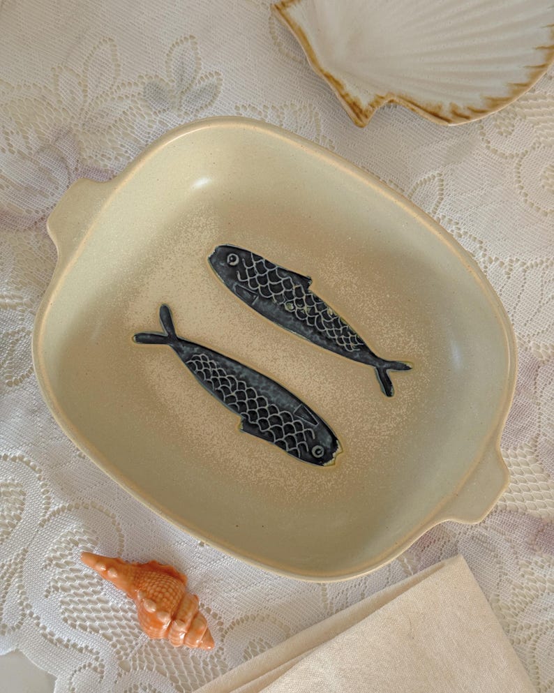 May include: A beige ceramic serving dish with two stylized blue fish designs. The dish has a square shape with rounded corners and small handles. A seashell and a napkin are also visible.