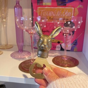 May include: A gold-colored bottle opener with a rabbit head design. The opener is held in front of a shelf with pink champagne glasses and a book titled "Taschen's New York". The background includes other decorative items.