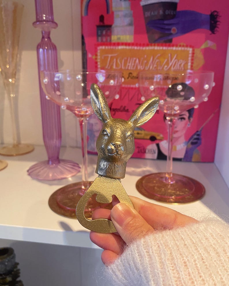 May include: A gold-colored bottle opener with a rabbit head design. The opener is held in front of a shelf with pink champagne glasses and a book titled "Taschen's New York". The background includes other decorative items.