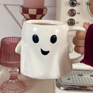 May include: A white ceramic ghost-shaped mug with black eyes and a smiling mouth, filled with a dark liquid. The mug has a handle and is being held. The background includes a coffee machine and a pink candle.