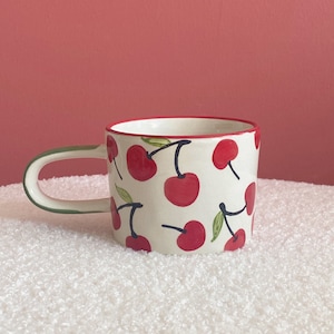 May include: A white ceramic mug with a red and green cherry pattern. The mug has a rounded handle.