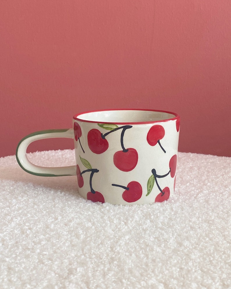May include: A white ceramic mug with a red and green cherry pattern. The mug has a rounded handle.