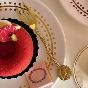 Può includere: Una forchetta da dessert color oro è appoggiata su un piatto bianco con pois dorati. Un dessert rosso con dettagli bianchi e verdi è posto su una base di carta nera. Una barretta di cioccolato rosa con un bordo decorativo è anch'essa sul piatto.