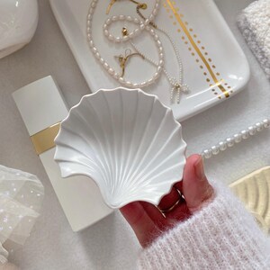 May include: A white, shell-shaped ceramic dish is held in a hand, with a white perfume bottle and a tray with jewelry in the background. The tray has gold accents and holds a pearl necklace, earrings, and a bracelet.