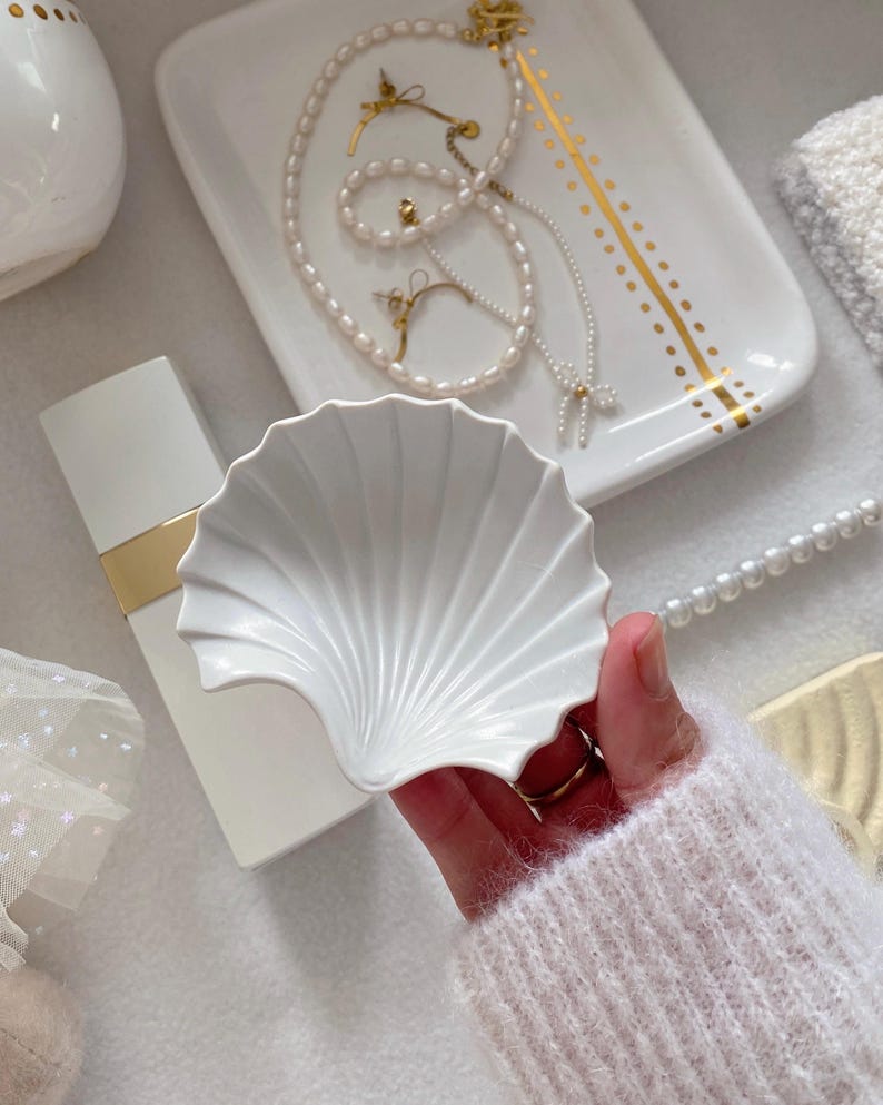 May include: A white, shell-shaped ceramic dish is held in a hand, with a white perfume bottle and a tray with jewelry in the background. The tray has gold accents and holds a pearl necklace, earrings, and a bracelet.