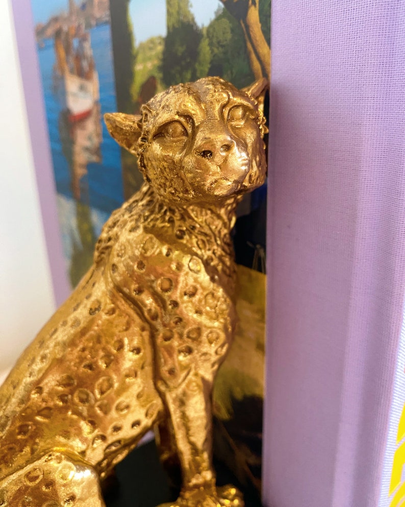 K&ouml;nnte beinhalten: Eine goldfarbene Gepardenfigur mit detaillierten Flecken und Gesichtsz&uuml;gen ist neben einem Buch mit lila Einband positioniert. Der Gepard scheint aus Metall oder einem &auml;hnlichen Material zu bestehen. Der Hintergrund zeigt ein Gem&auml;lde mit einer K&uuml;stenlandschaft.
