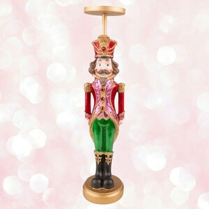 May include: A gold candle holder shaped like a traditional Christmas nutcracker. The nutcracker is wearing a red and green uniform with gold accents. The candle holder is approximately 12 inches tall.