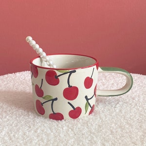 May include: A white ceramic mug with a green handle features a red cherry pattern. The mug is sitting on a white fluffy surface.
