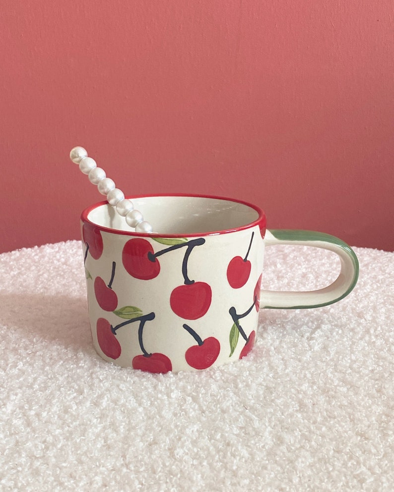 May include: A white ceramic mug with a green handle features a red cherry pattern. The mug is sitting on a white fluffy surface.