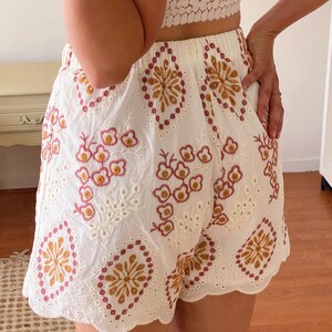 K&ouml;nnte beinhalten: Wei&szlig;e Baumwollshorts mit einem gewellten Saum und einem gestickten Blumenmuster in den Farben Rosa, Orange und Braun.