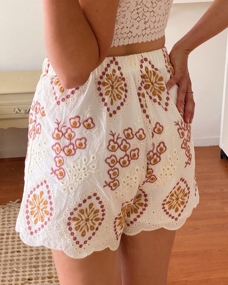 K&ouml;nnte beinhalten: Wei&szlig;e Baumwollshorts mit einem gewellten Saum und einem gestickten Blumenmuster in den Farben Rosa, Orange und Braun.