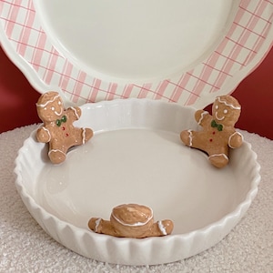 Puede incluir: Un plato de cerámica blanco con borde festoneado, con tres figuras de hombre de jengibre. El plato de atrás tiene un patrón de cuadros rosas. Los hombres de jengibre tienen detalles de glaseado blanco y acentos rojos y verdes.