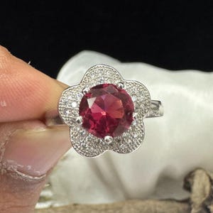 May include: A silver ring featuring a large, round, deep red gemstone at the center, surrounded by smaller, clear stones in a flower-shaped setting. The ring is held up against a white cloth background.