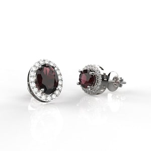 May include: A pair of stud earrings featuring oval, deep red gemstones surrounded by a halo of smaller, clear stones. The earrings are set in a silver-colored metal, with a post and butterfly backing.
