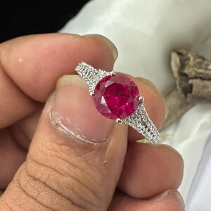 Puede incluir: Un anillo de plata con una gran gema roja redonda y brillante. La banda está adornada con piedras más pequeñas y claras, añadiendo brillo. El anillo se sostiene, mostrando su diseño y color.