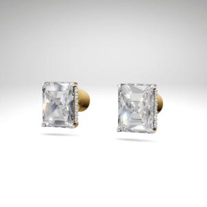 May include: A pair of square-cut, clear gemstone stud earrings. Each earring has a gold-colored setting with a halo of small, clear stones around the main stone. The earring posts are also gold-colored.